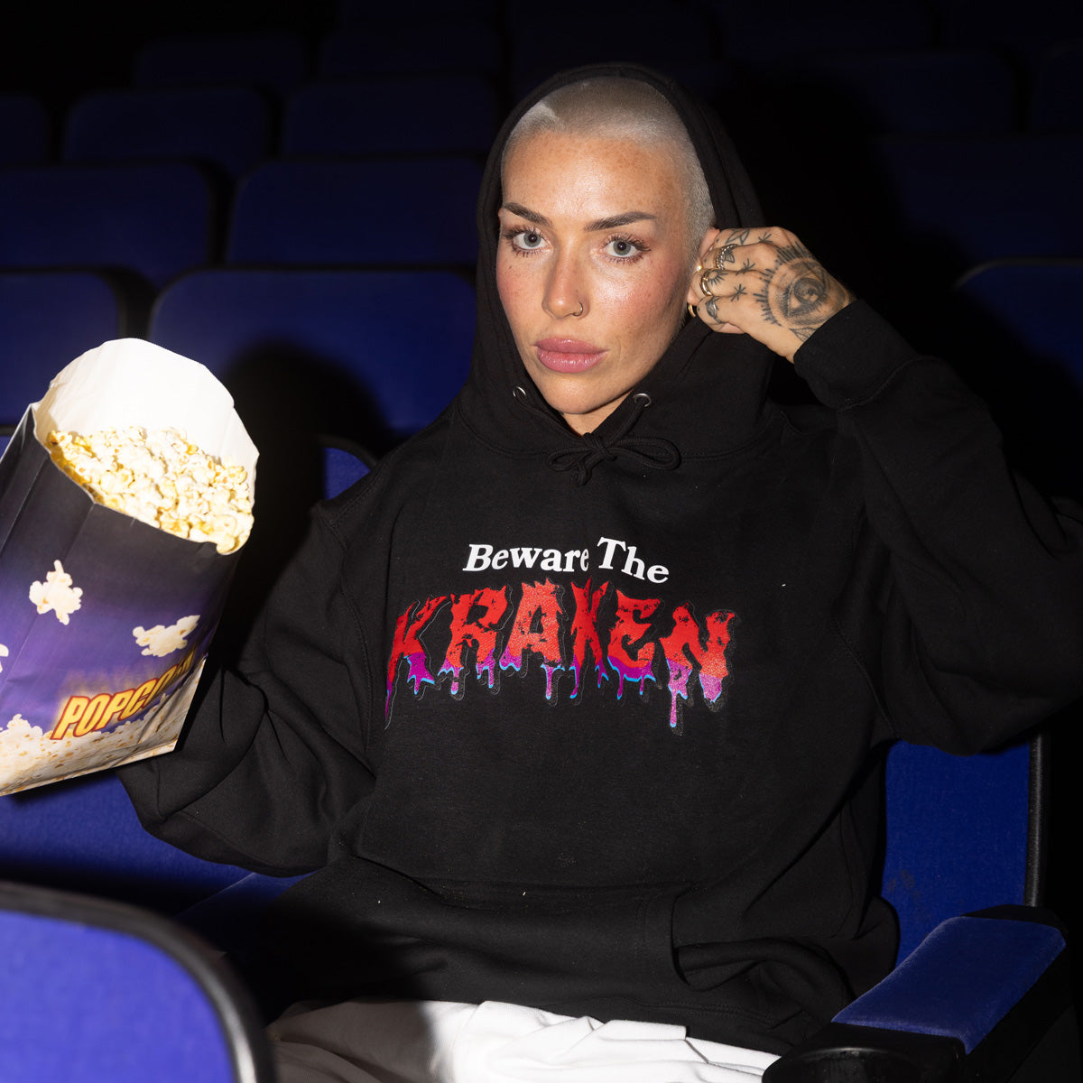 Front image of Beware The Kraken Hoodie in black with red and purple drip design of "KRAKEN" in the middle on a female model in a dark and empty  movie theater holding a bag of popcorn