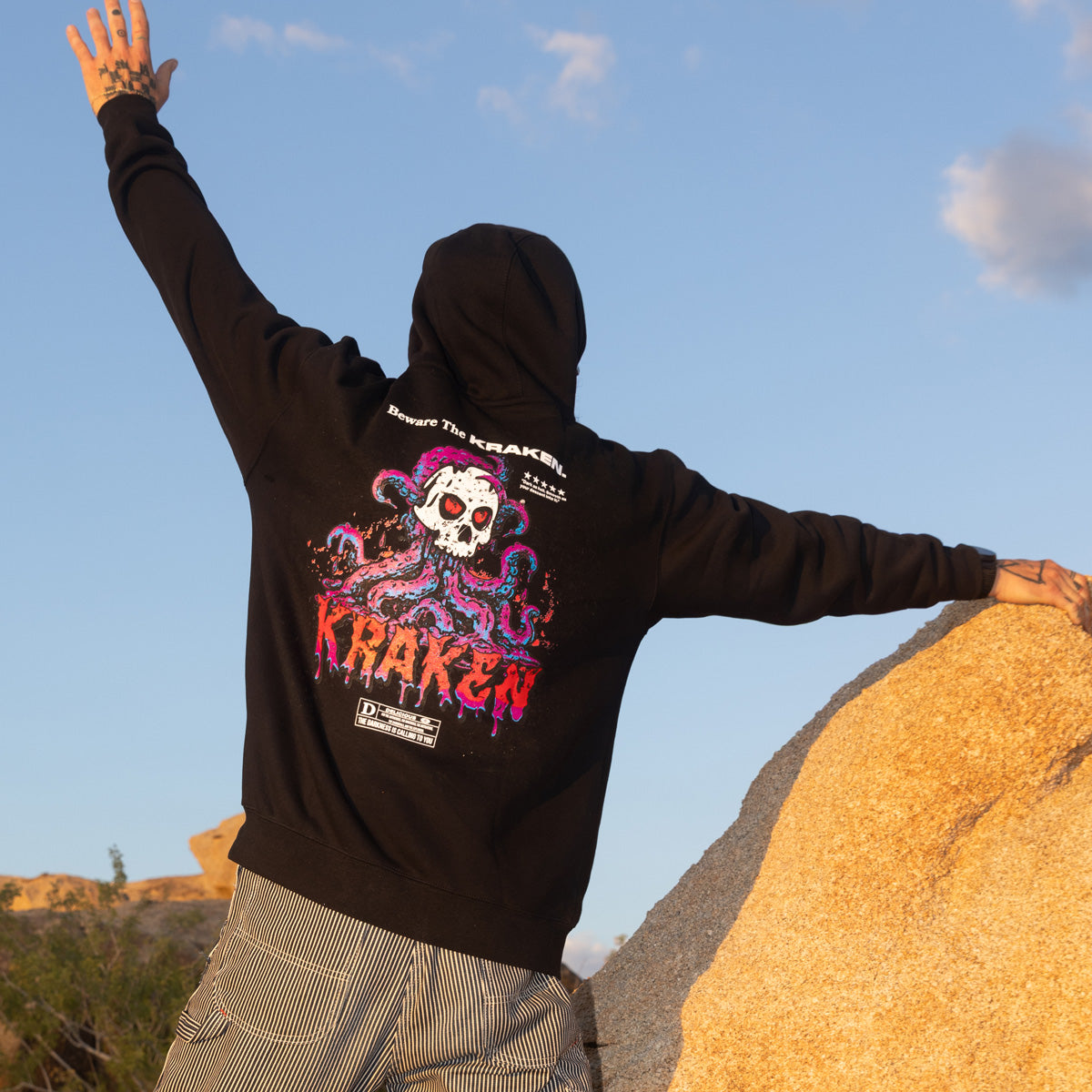 Back view of the Beware The Kraken Hoodie in black with a graphic of a white skull with red eyes surrounded by purple and teal kraken tentacles, with dripping red text that reads 'KRAKEN' on a male model climbing boulders in the desert
