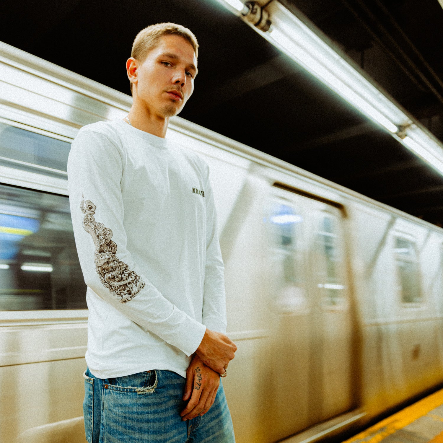 The image depicts a person standing on a subway platform in an urban setting wearing the Artist of the Abyss long sleeve.