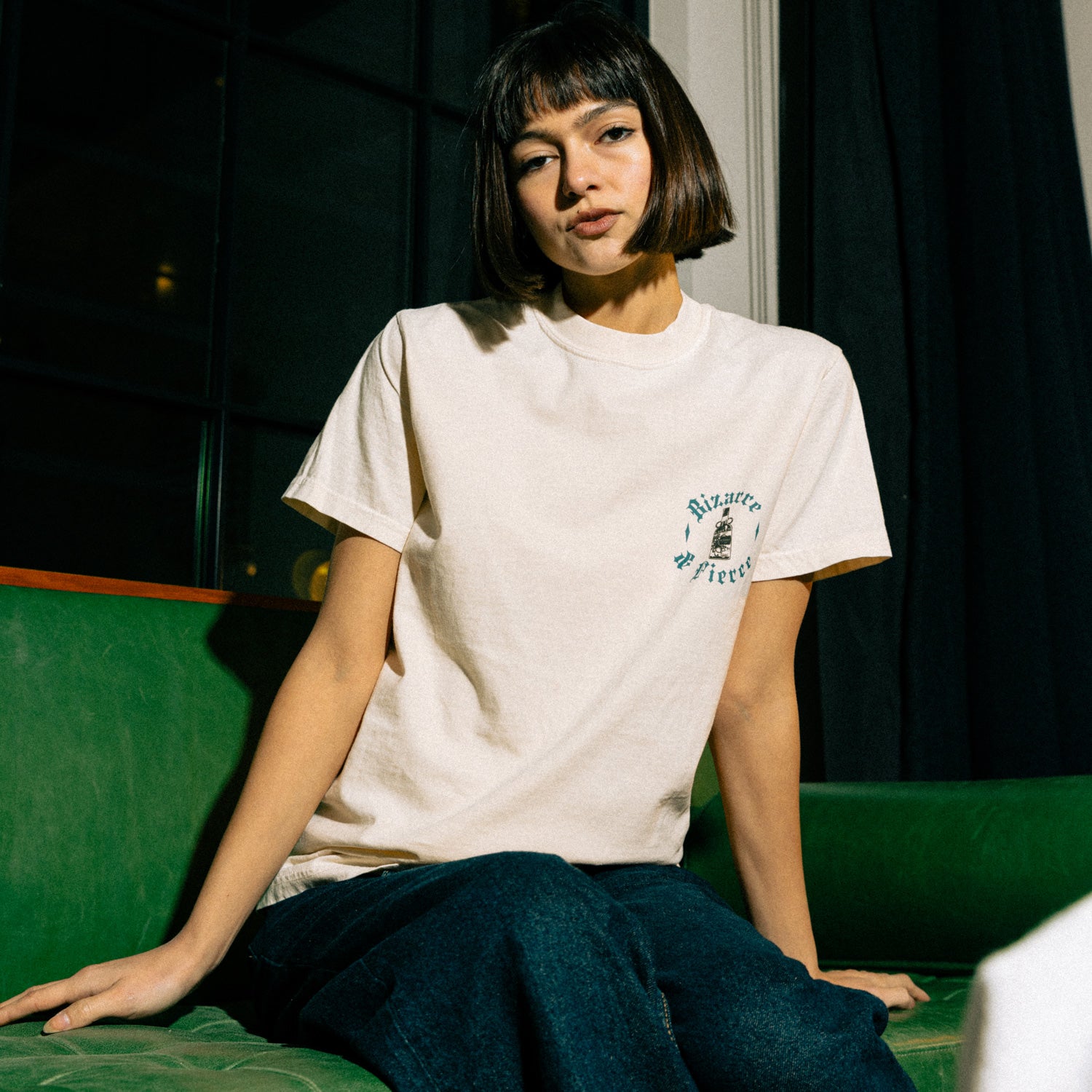 The image depicts a person seated on a green couch in a dimly lit room with large windows and dark curtains. The tee has a graphic on the left chest that reads "Bizarre & Fierce".