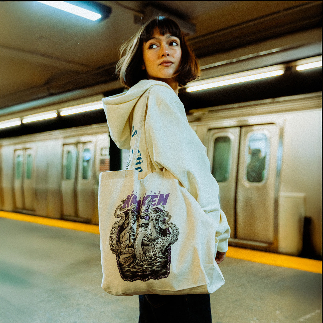The image depicts a person standing on a subway platform in an urban setting. The focus is on the tote bag the person is carrying, which features the Artist of the Abyss design.