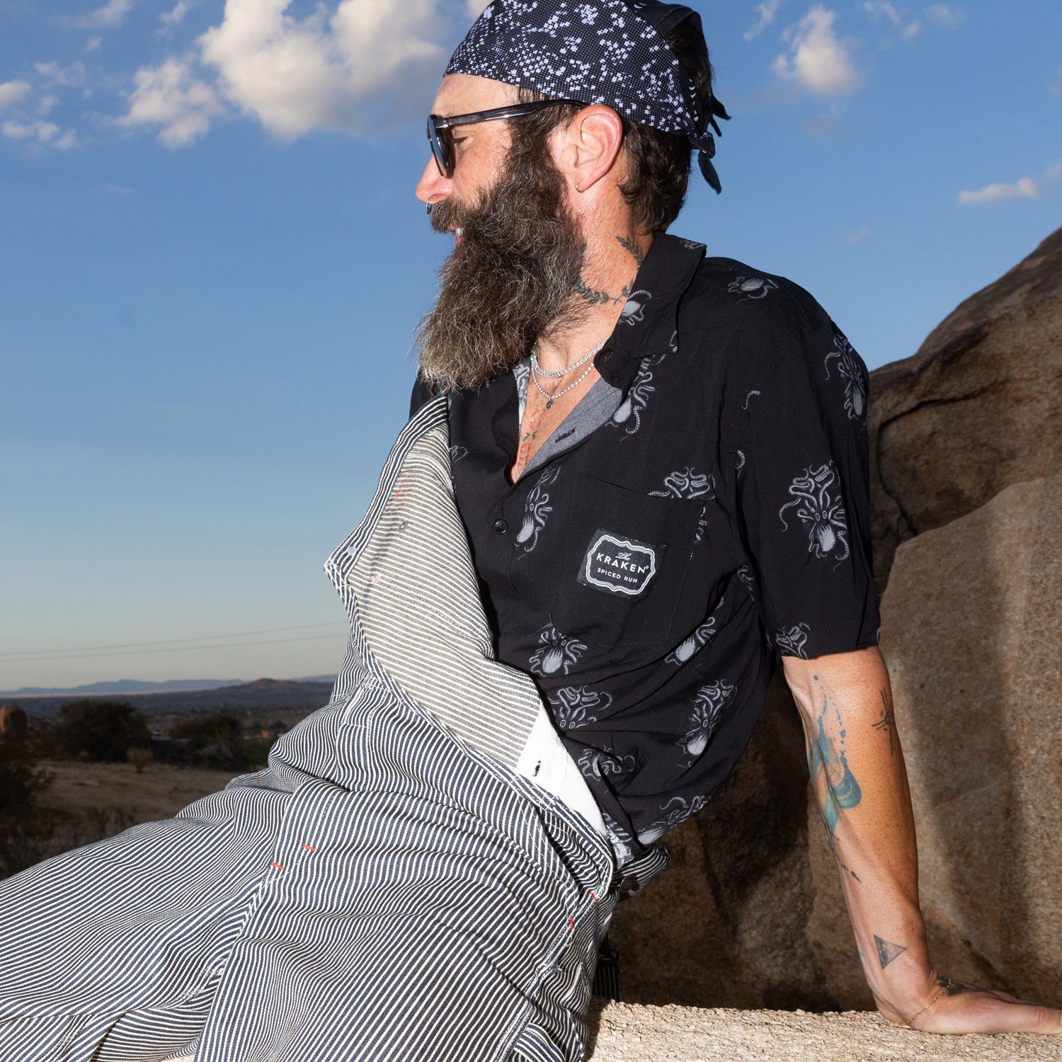 Lifestyle image of the Button Down shirt on a male model. The mode is bearded and styled with striped coveralls and a black and white bandana and black sunglasses.