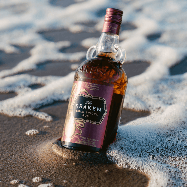 A Kraken Gold Spiced Rum bottle on a sandy beach with the sea in the background.