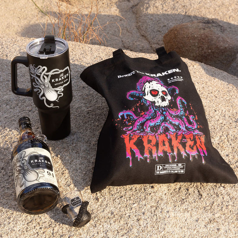 Image of Kraken merch and black spiced rum laid out on a boulder in the desert. The items are The Kraken Black Spiced Rum, Kraken Tumbler, Kraken Shot Glass, and Beware The Kraken Tote bag.