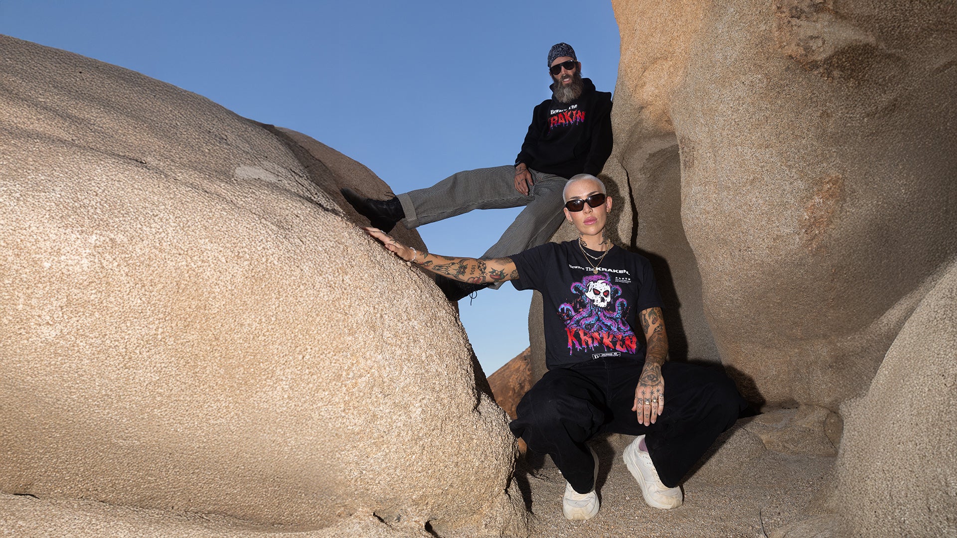 Lifestyle image of two models wearing Beware The Kraken merch posing between boulders in the desert. The male model is wearing the Beware The Kraken Hoodie in black and the female model is wearing the Beware The Kraken tee in black.