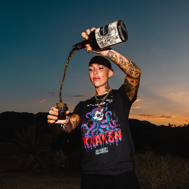 Image of female model pouring Kraken Black Spiced Rum into shot glass in the desert wearing Beware The Kraken tee in black