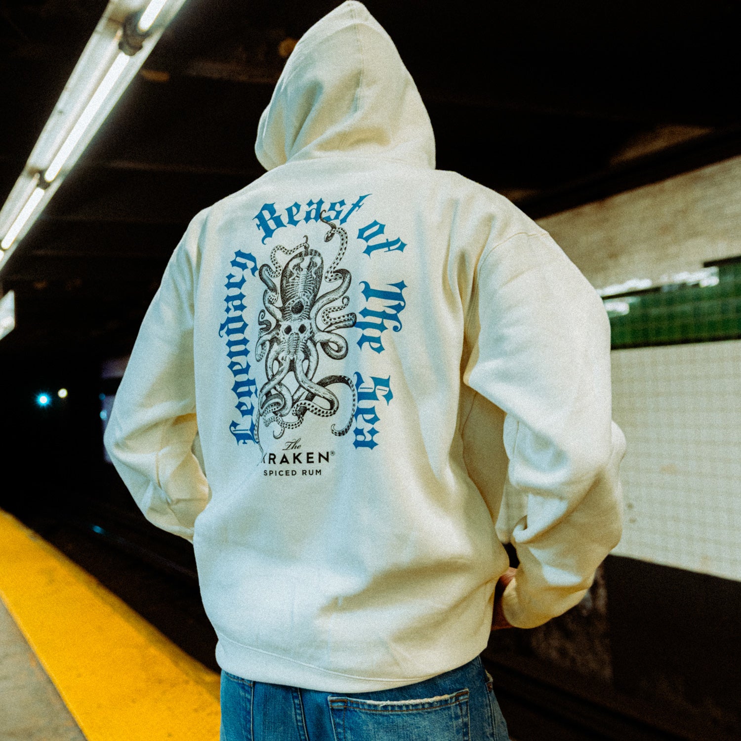 The image depicts a person standing on a subway platform wearing a white hoodie featuring a distinctive graphic design on the back. The design prominently includes The Kraken illustration and the text "Legendary Beast of the Sea" with The Kraken logo.