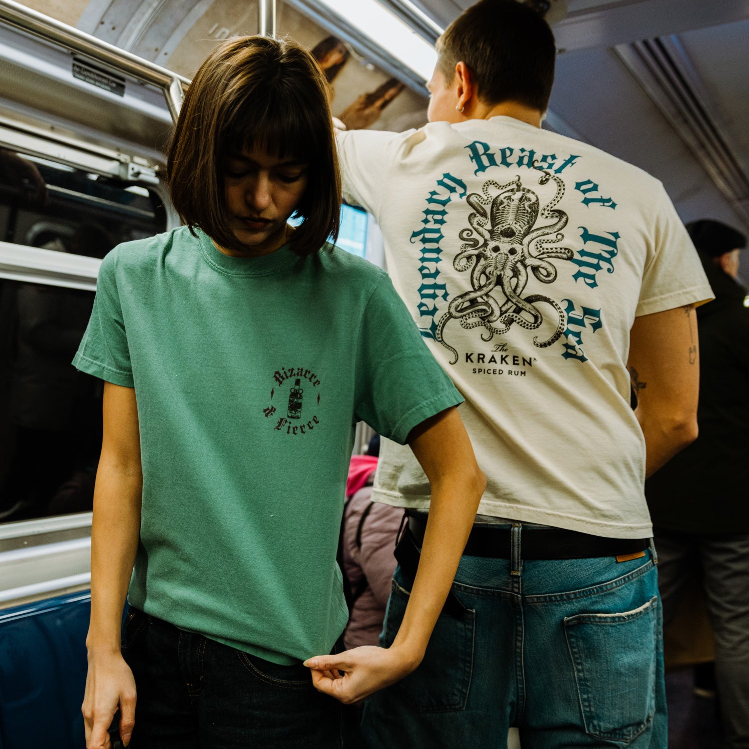 This image shows two models wearing the Legendary Beast of the Sea tee, one in green, one in ivory in a subway setting.