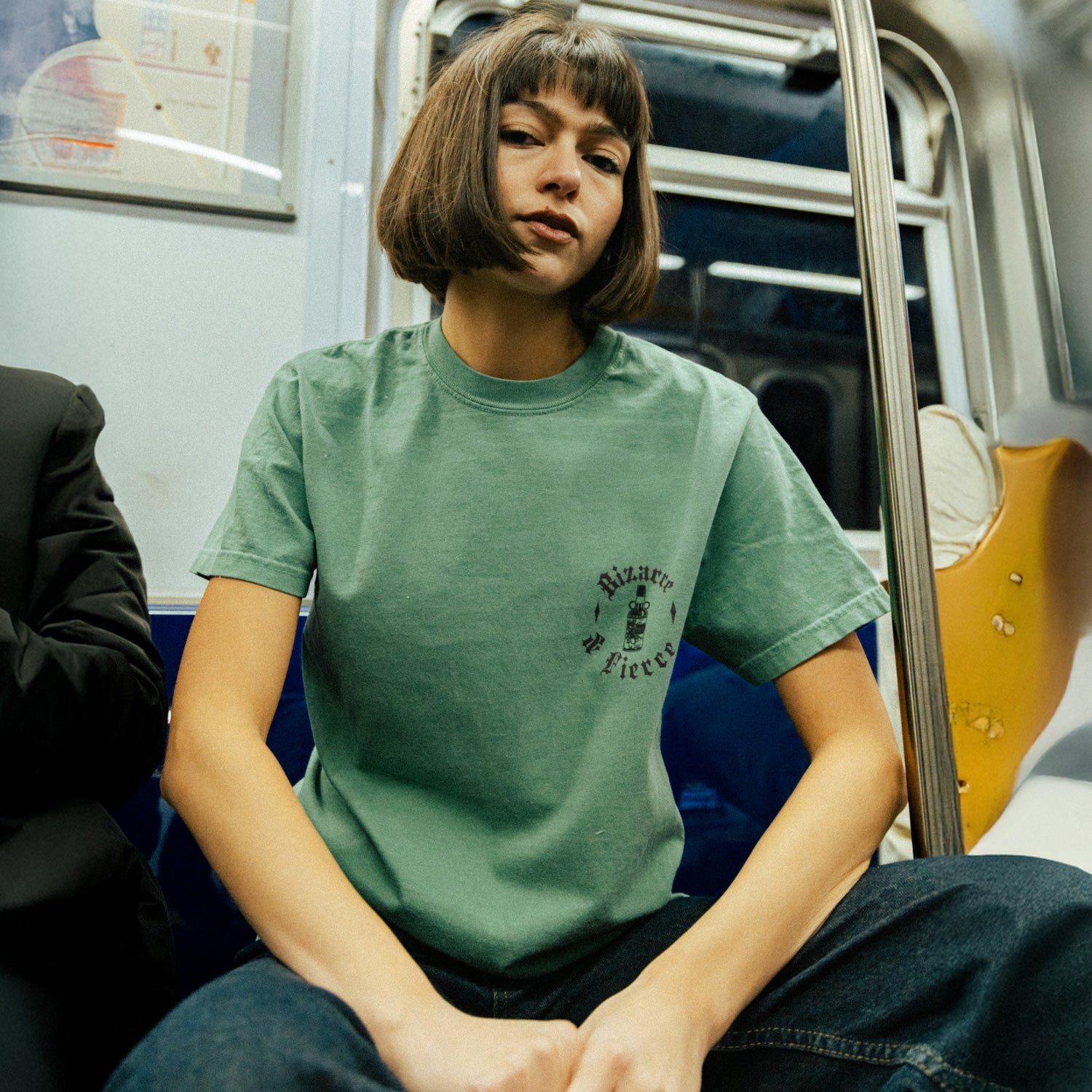 The image depicts a person sitting on a subway train, wearing a green T-shirt with a small graphic and text "Bizarre & Fierce" on the left chest area. The setting in an urban public transit environment.