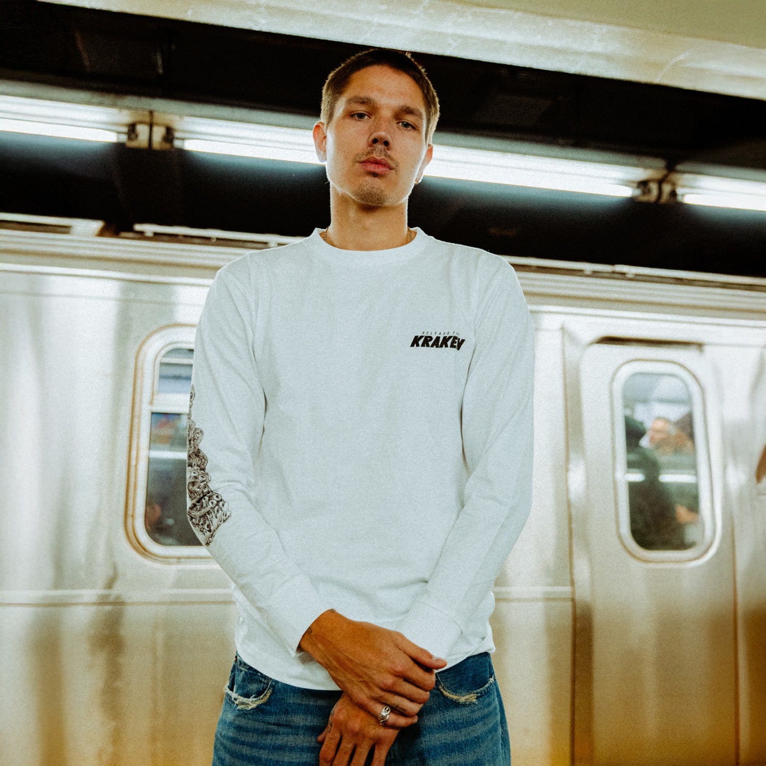 The image depicts a person standing on a subway platform in an urban setting wearing the Artist of the Abyss long sleeve.