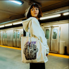 The image depicts a person standing on a subway platform in an urban setting. The focus is on the tote bag the person is carrying, which features the Artist of the Abyss design.