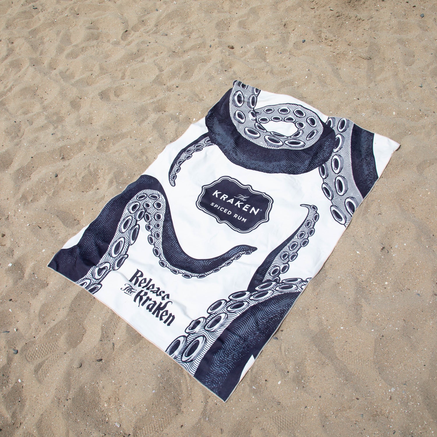 A white beach towel with The Kraken tentacle print and 'Kraken Spiced Rum' branding, laid flat on a sandy beach.